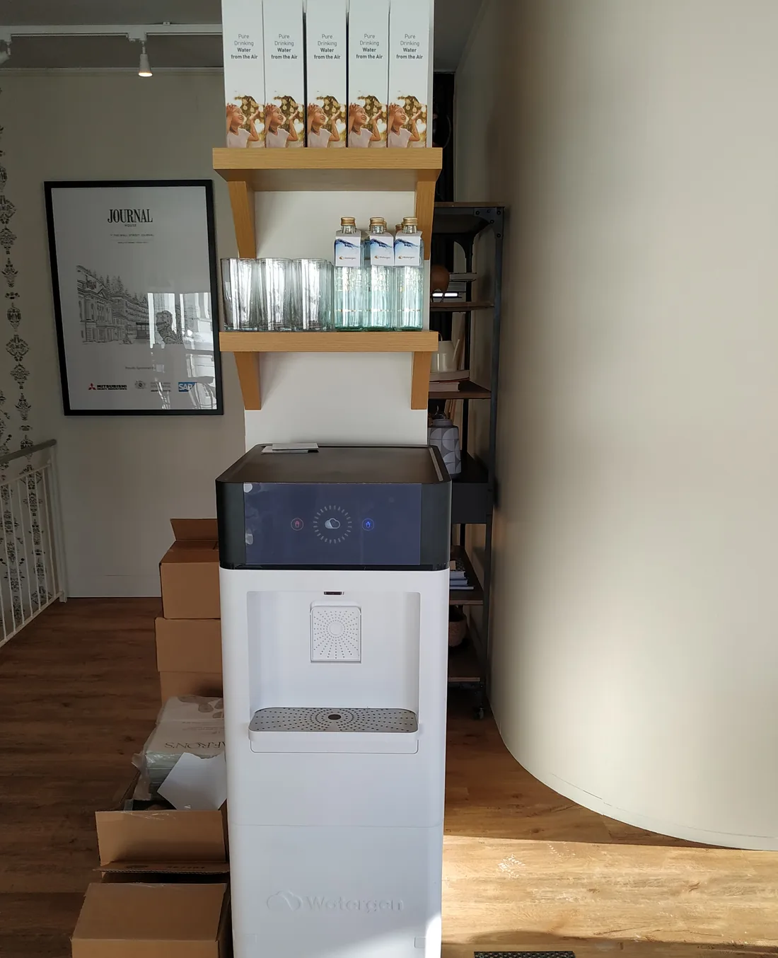 A Watergen generator at Journal House, with glass bottles and boxes reading “Pure Drinking Water from the Air” on the shelf above