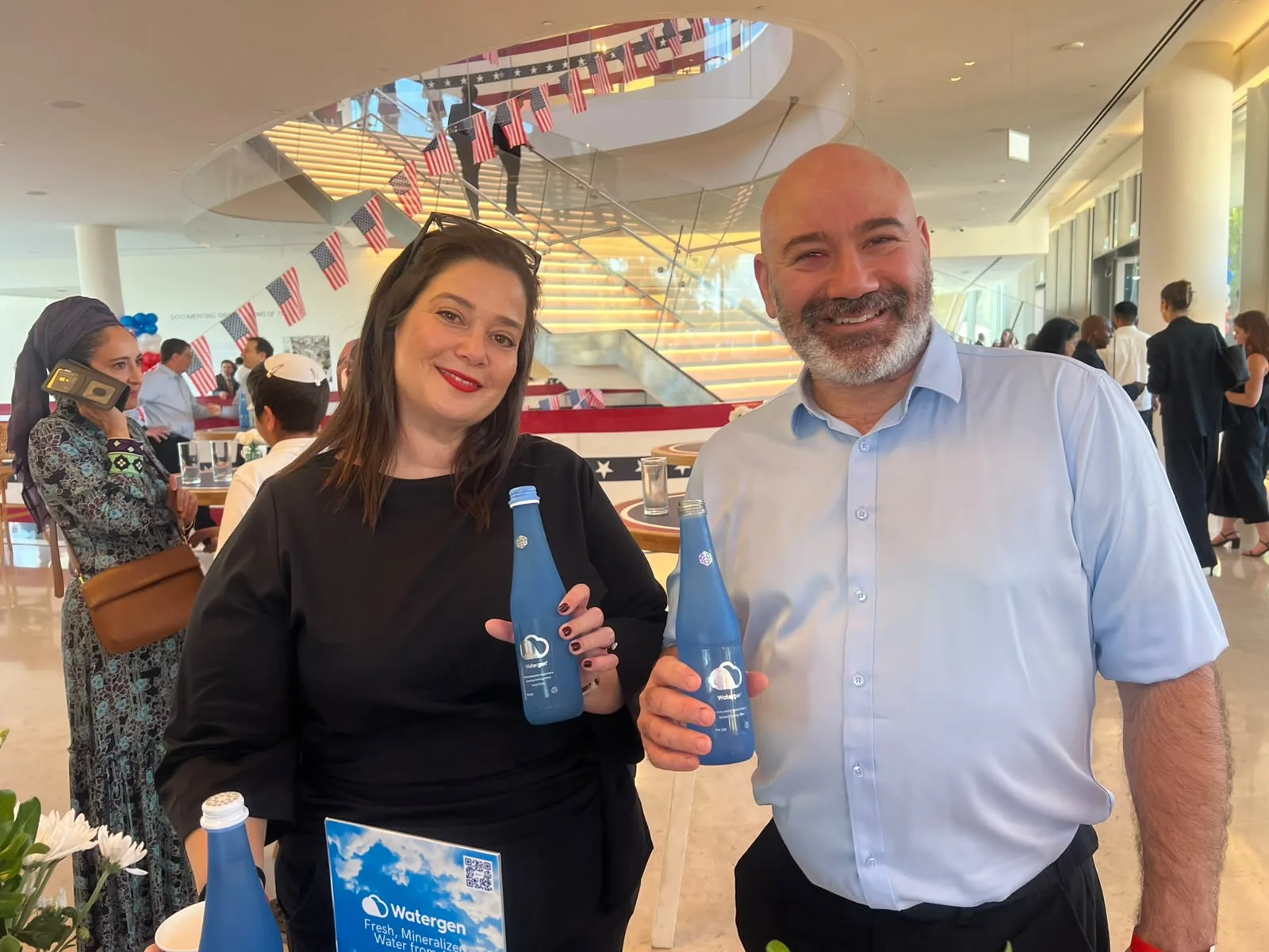 Two guests holding blue Watergen bottles at the celebration, a staircase strung with American flags behind them