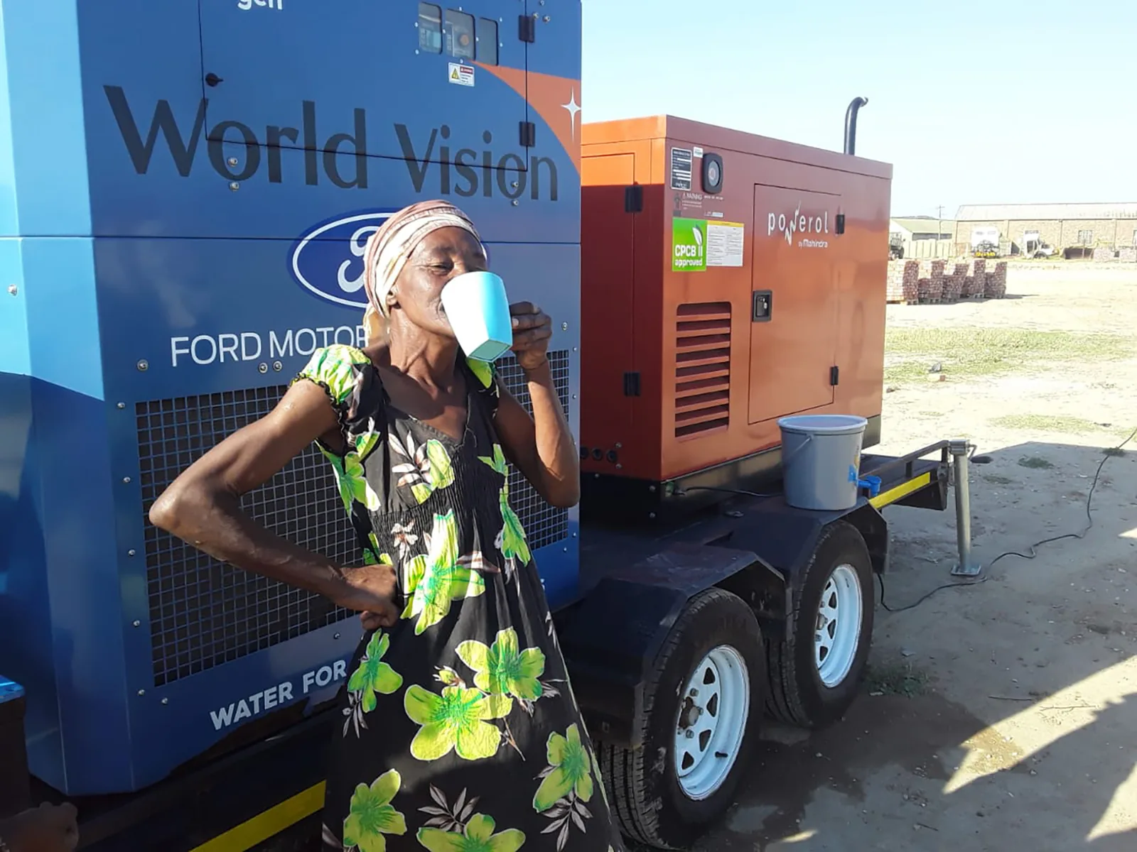 A woman drinking water beside a trailer-mounted Watergen unit marked World Vision