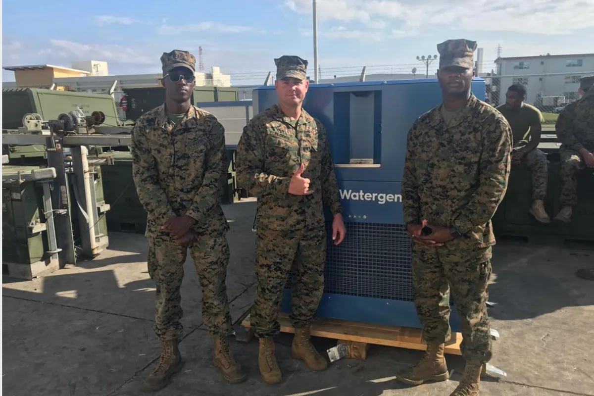 Soldiers standing beside a Watergen atmospheric water generator at a military base